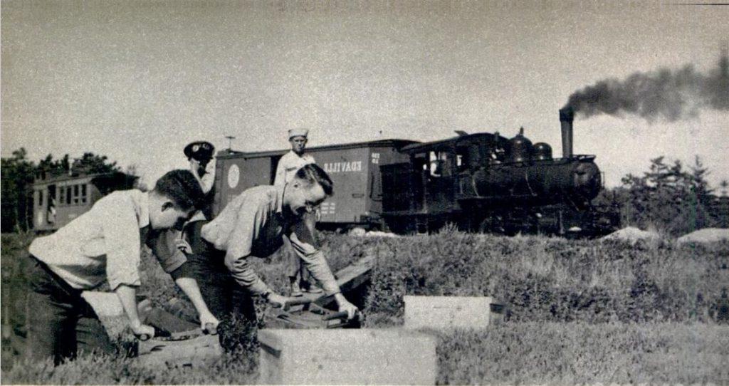 Black and white photograph of workers in a field with a train passing behind them. In the foreground, two kneeling workers are bending over and holding onto wooden crates. Two other workers stand behind them and watch. The train in the background has smoke billowing out of its chimney and the word “Edaville” written on its side.